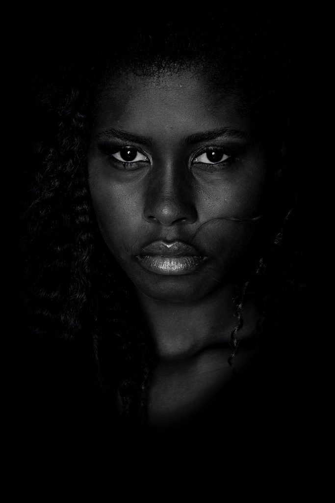 Image of a Black woman on a black background; her expression is serious, looking straight at the camera.
